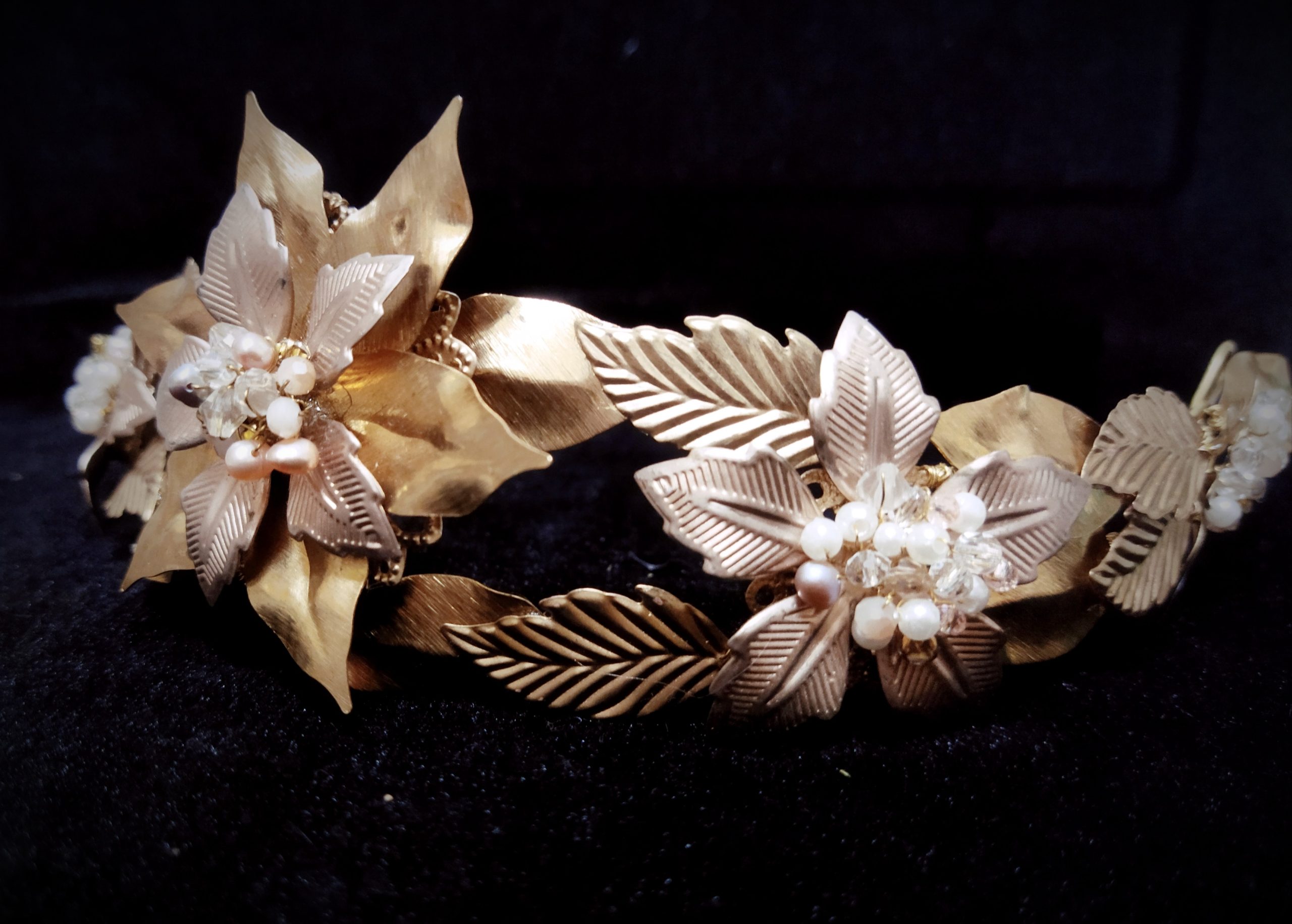 Tocado artesanal de hojas doradas y nacaradas con flores y perlas sobre fondo oscuro.